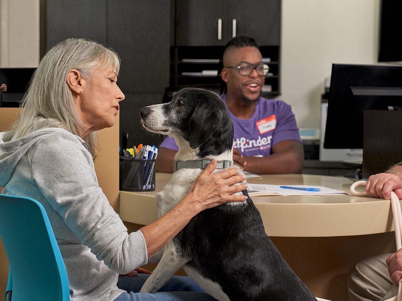 Person holding a black-and-white dog while talking with staff at a Petfinder adoption center.